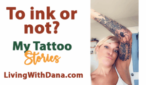 A woman playfully lifts her arm to show detailed tattoos on her inner arm, including a spider and floral designs. She’s smiling and expressive in a white tank top indoors.