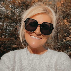 A woman smiling in sunglasses, with a casual tank top, outdoors in a sunny setting with trees and a lake in the background.